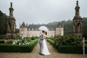 les mariés s’embrassent devant un chateau, sous un parapluie transparent, photo de couple sous la pluie