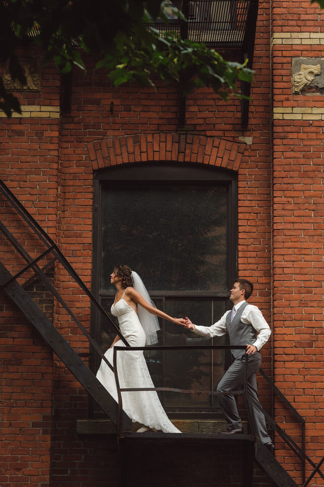 Mariés se tenant les mains en montant des escaliers, Séance photos de mariage dans un espace urbain avec un mur en brique rouge, Mariage à Montréal à l'Espace Canal, plan B